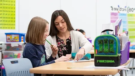 Student teacher helping elementary school student at her desk.