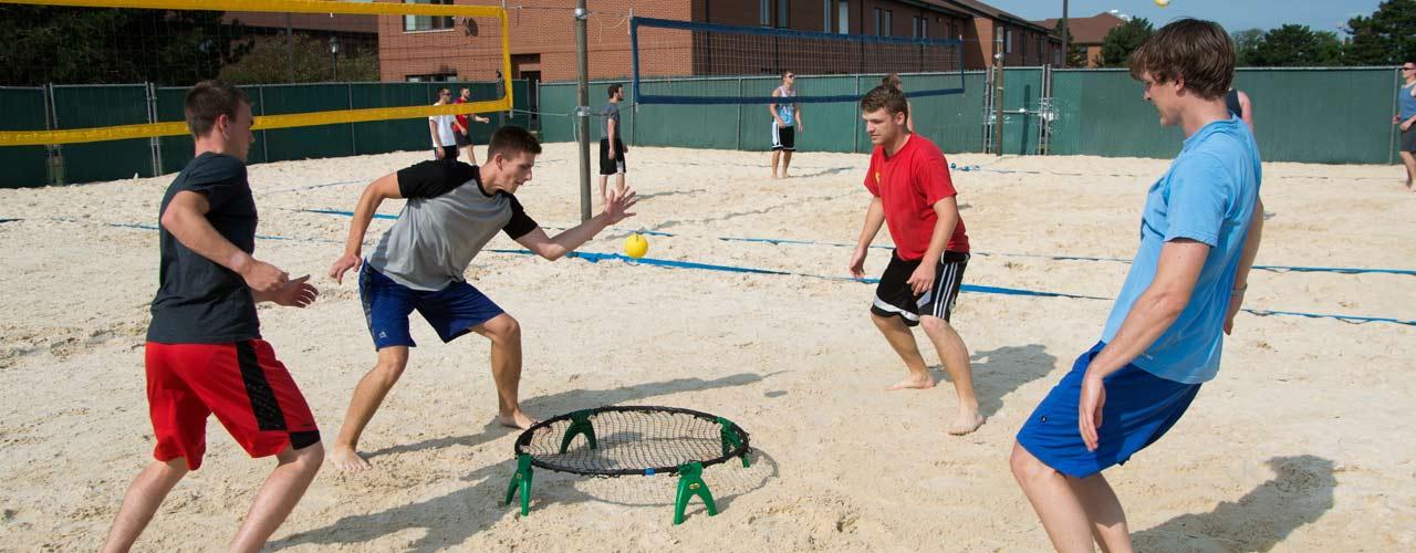 A group of male students play spikeball in a sand volleyball court
