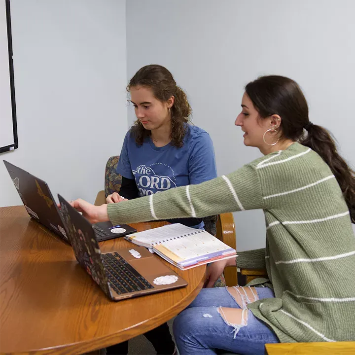 女学生导师坐在桌子旁帮助另一名女学生使用笔记本电脑和教科书.