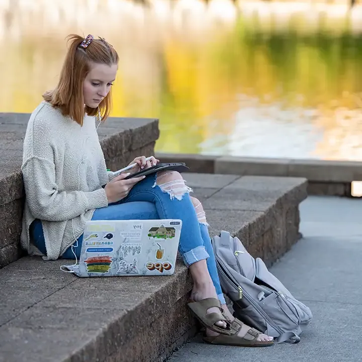 Woman sitting on outside steps by a pond working on laptop